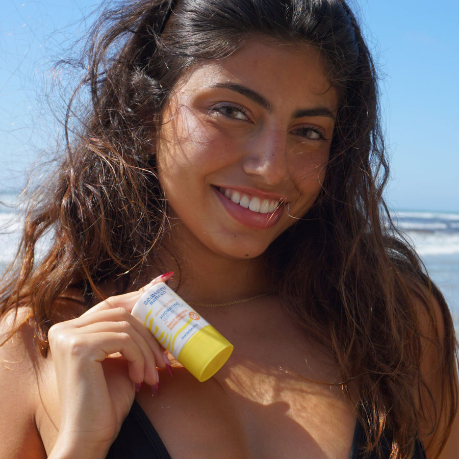 Dark skinned, brunette woman at the beach in a black bikini top holding Seaweed Bath Co. Refresh & Protect Hydrating Serum Stick SPF 50.