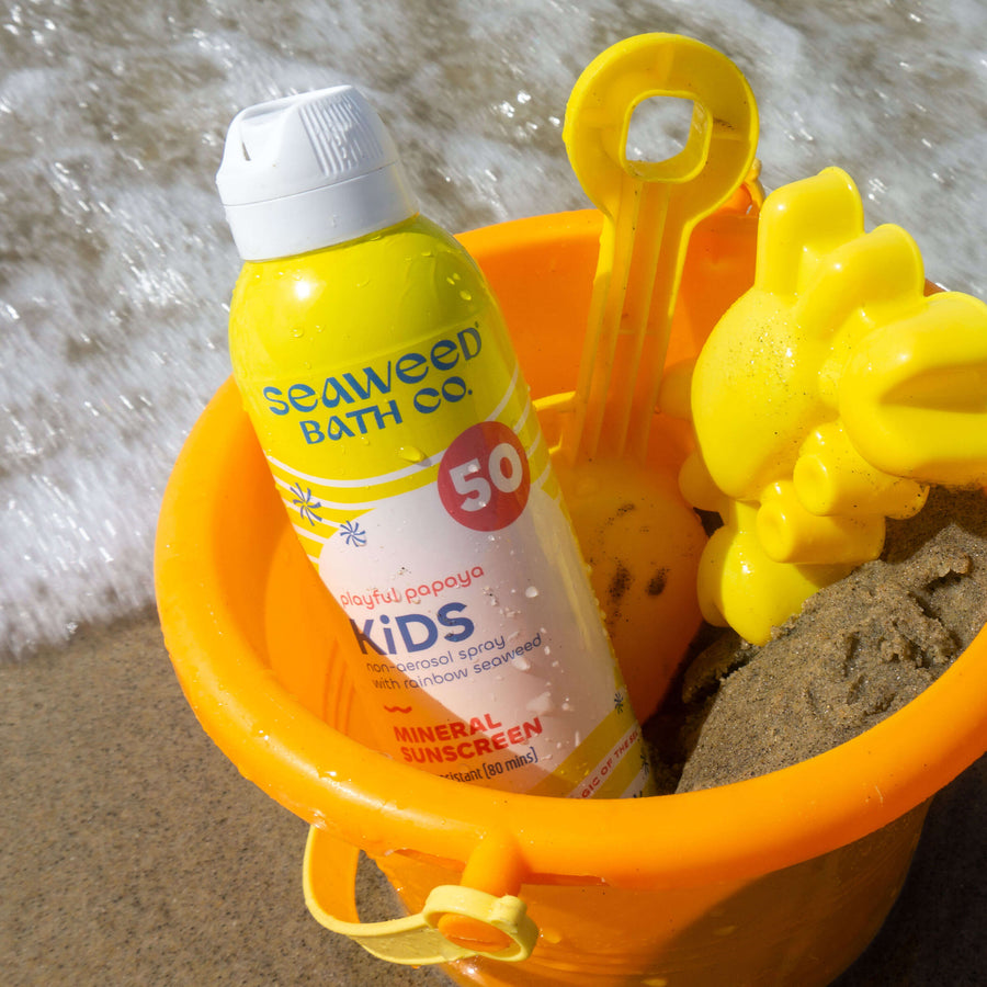 Kids Spray SPF 50 in a bucket with sand and yellow beach toys in the ocean surf.