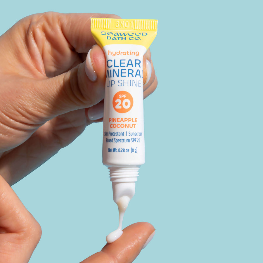 Hand squeezing Seaweed Bath Co. Hydrating Clear Mineral Lip Shine SPF 20 Tube with lip balm dripping onto finger.