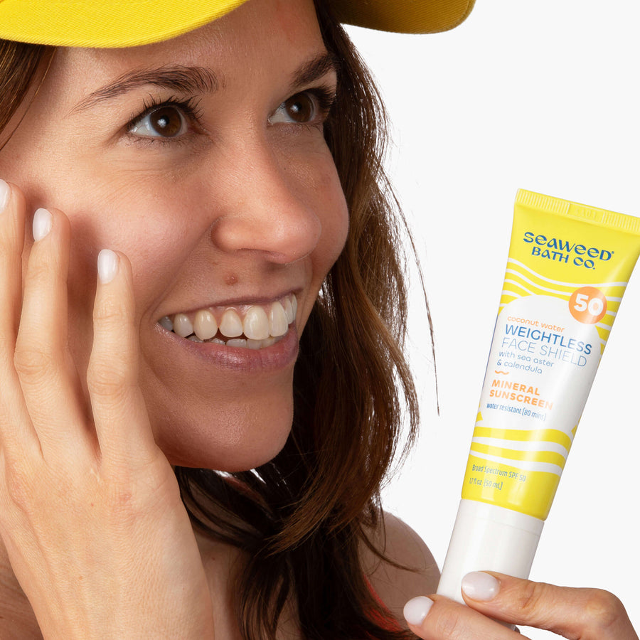 Brunette woman wearing yellow baseball  with fingers touching face and holding Weightless Face Shield SPF 50.