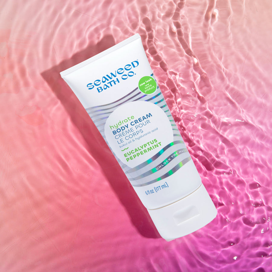 Seaweed Bath Co. Hydrate Body Cream in Eucalyptus Peppermint Scent tube laying in rippling water on sunset pink background.