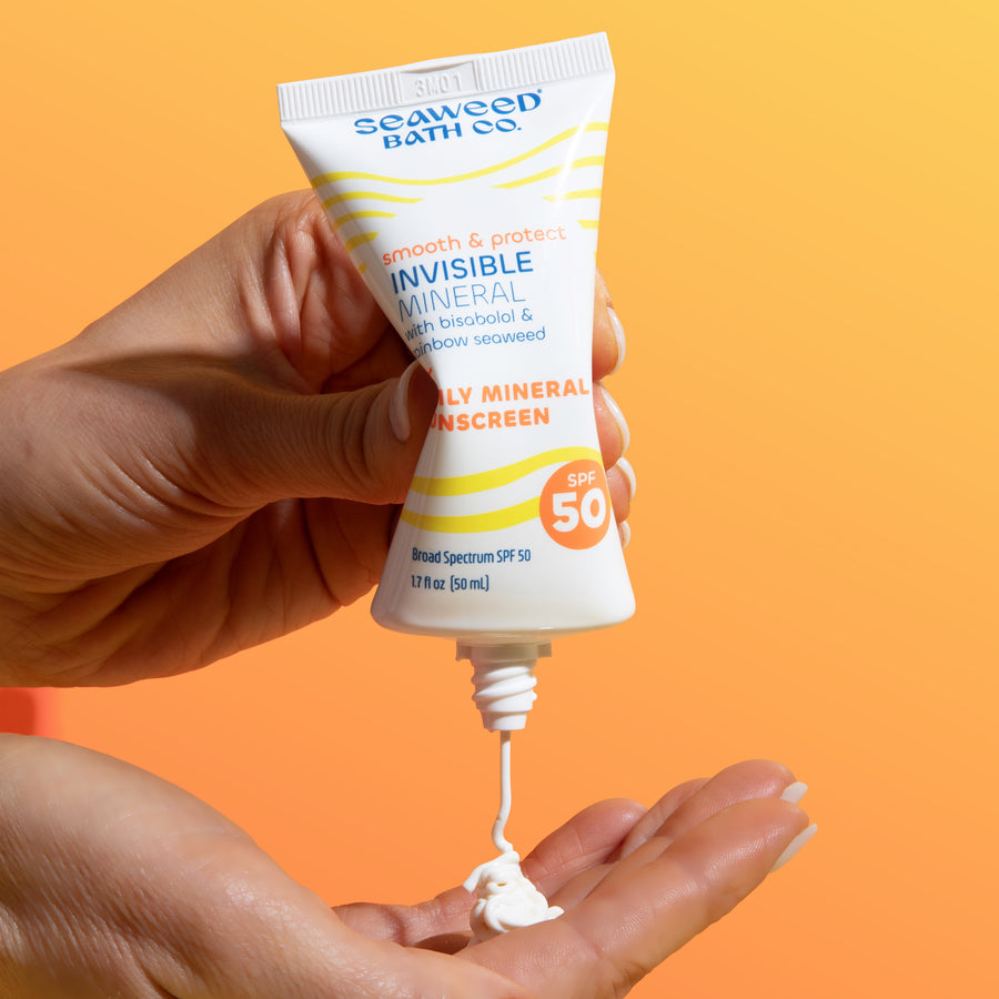 Hand squeezing Seaweed Bath Co. Invisible Mineral SPF 50 with white gel cream squirting onto another hand. On orange gradient background.
