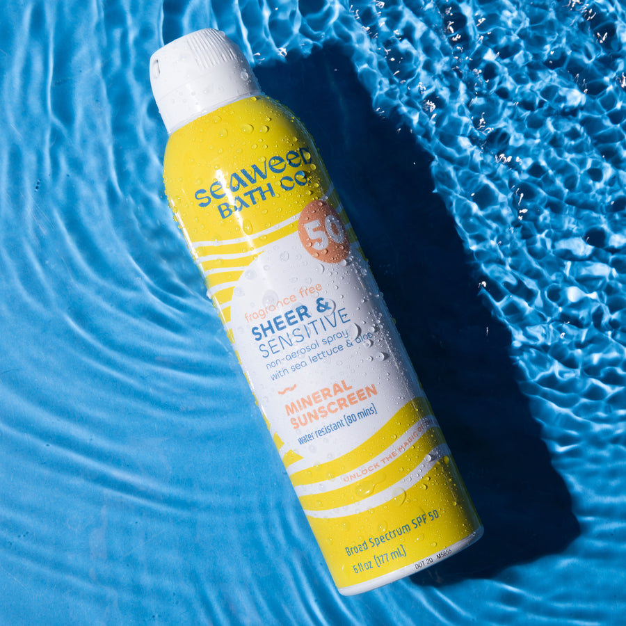 Sheer and Sensitive Spray SPF 50 Sunscreen laying flat in rippling water on sky blue background. Seaweed Bath Co.