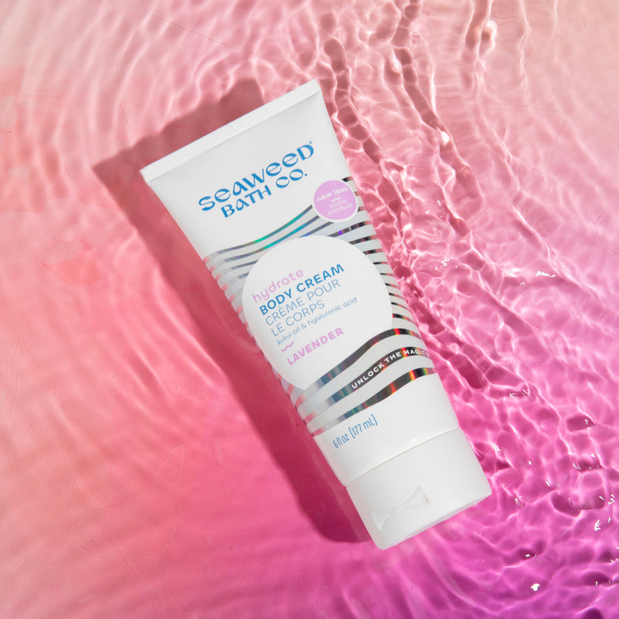 Hydrate Body Cream in Lavender tube laying in rippling water on sunset pink background. Seaweed Bath Co.