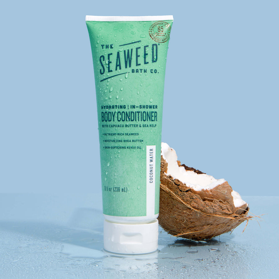 Seaweed Bath Co. Hydrating In-Shower Body Conditioner next to half a coconut on sky blue background
