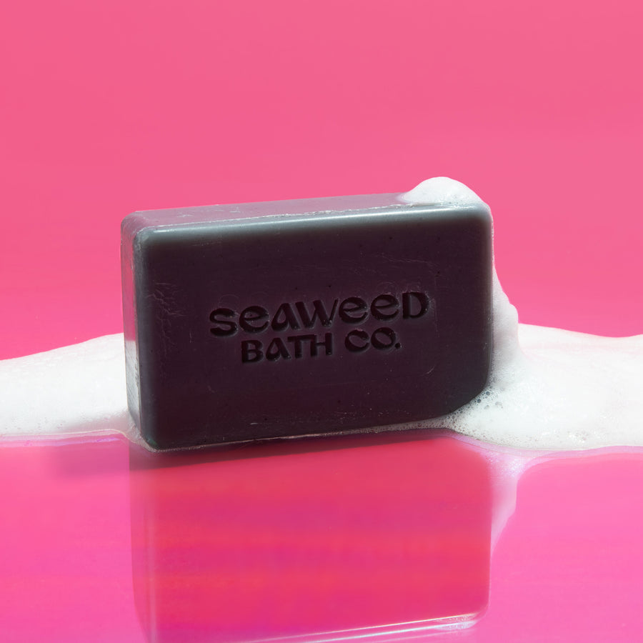 Seaweed Bath Co. Purifying Detox Facial Bar covered in soap bubbles on pink background.