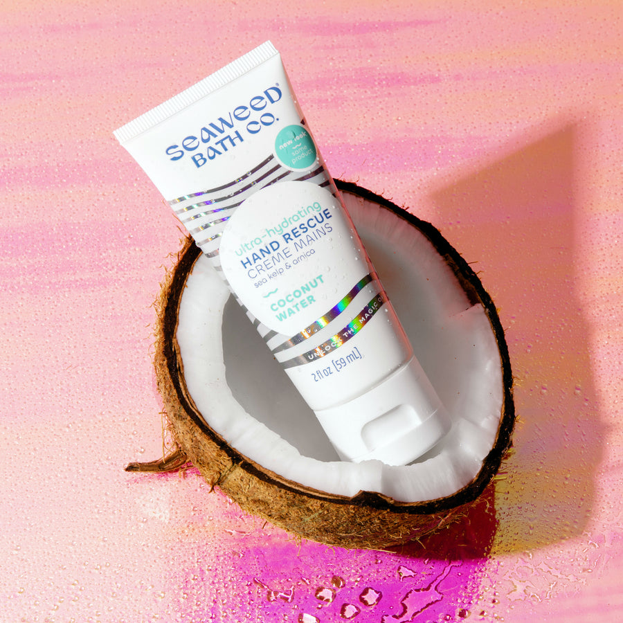 Seaweed Bath Co. Ultra-Hydrating Hand Rescue resting in a split coconut with water droplets on a holographic background.